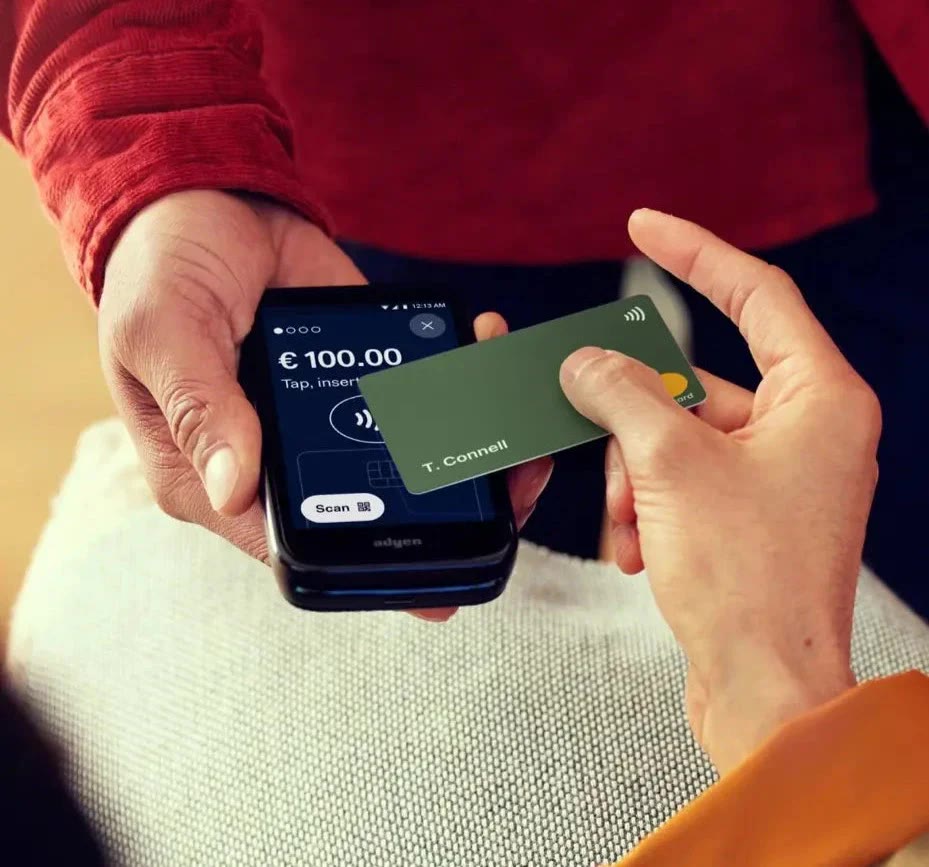 A shopper taps a card on an Adyen AMS1 held by store staff, showing €100.00